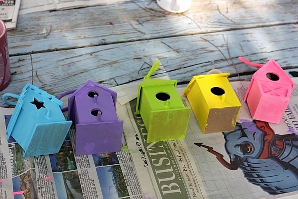 Five birdhouses painted with acrylic paint laying on newspaper drying 
