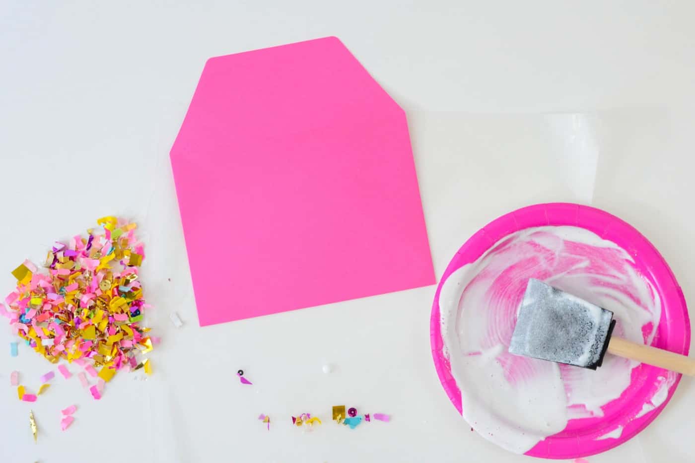 Pile of confetti, pink envelope, and a foam brush with Mod Podge on a plate
