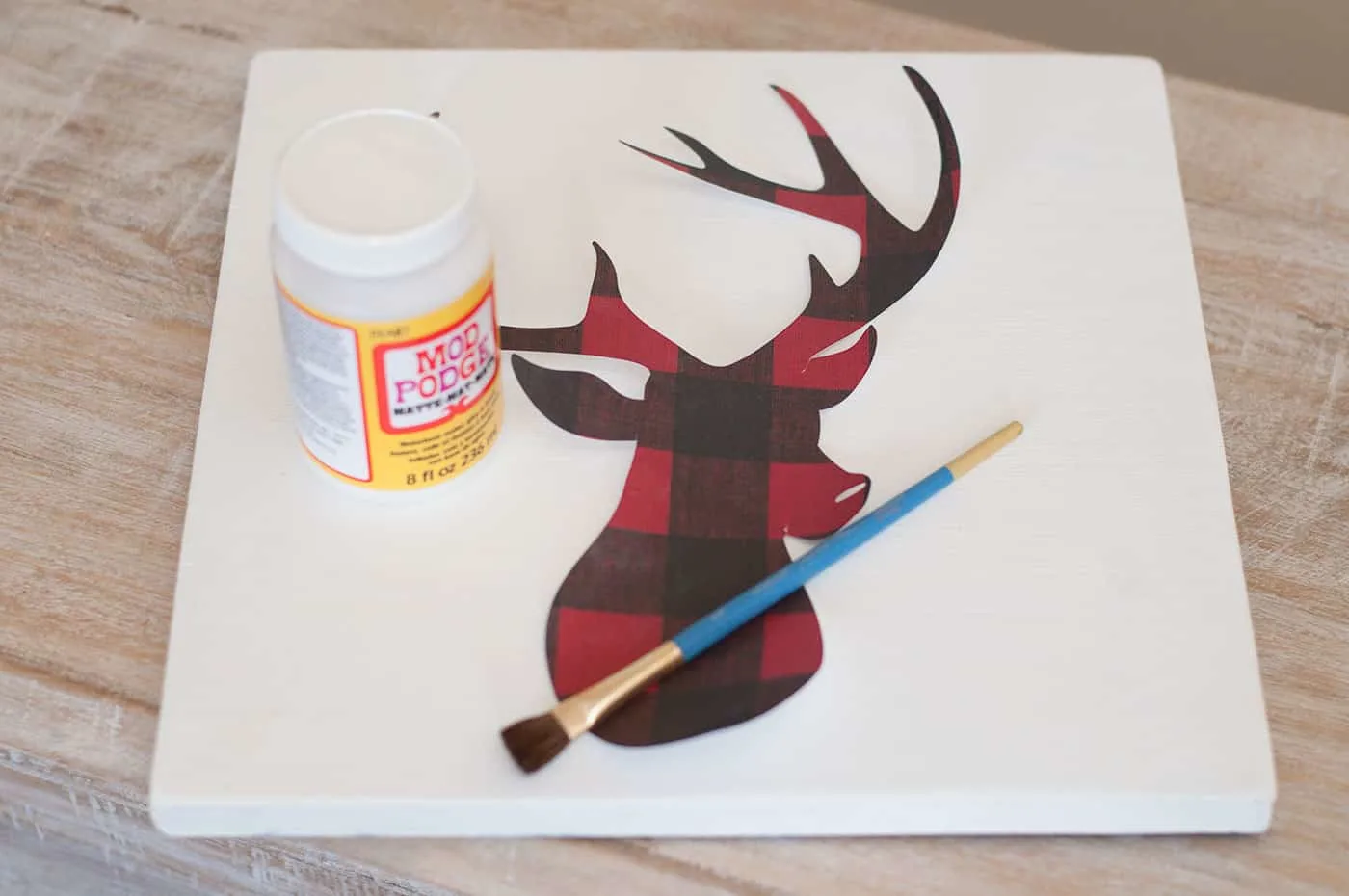 White wood piece, plaid deer head cut out of scrapbook paper, Mod Podge Matte, and a paintbrush