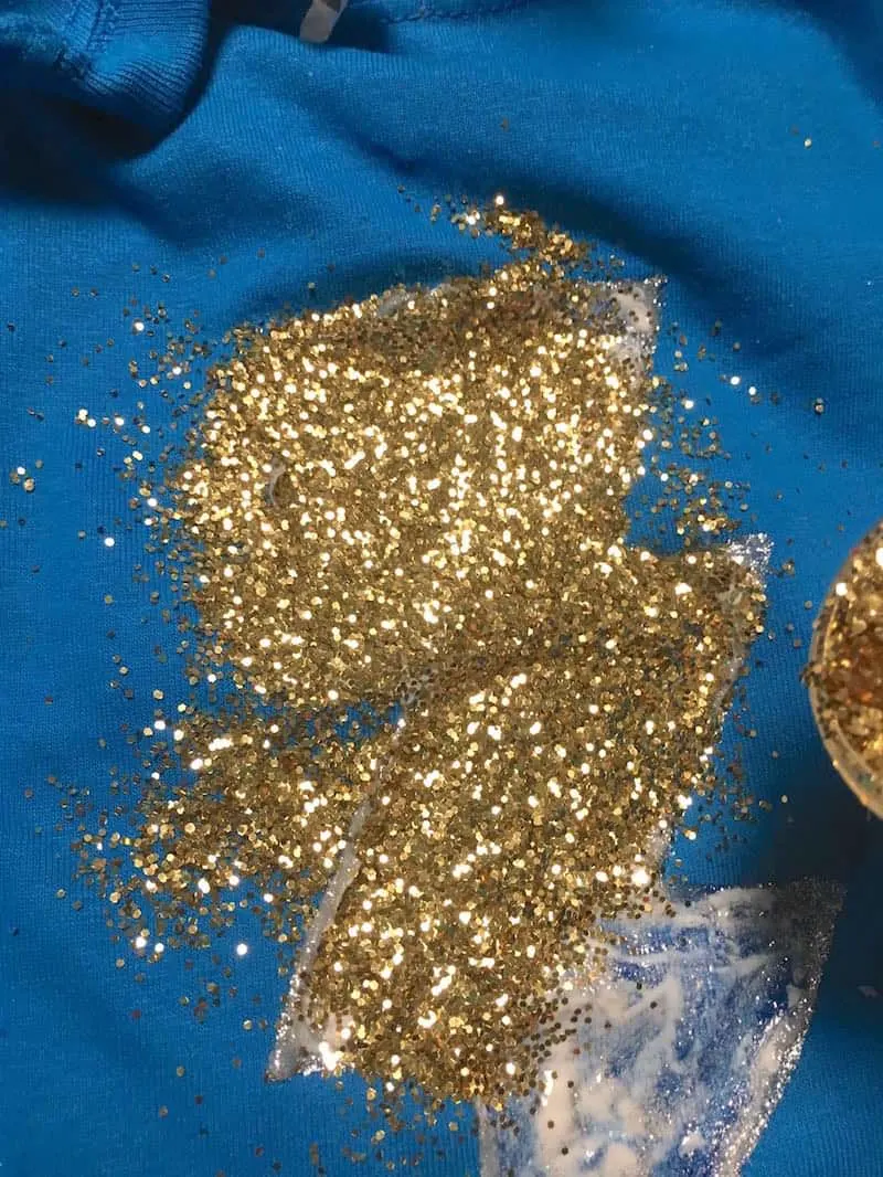Sprinkling gold glitter on Mod Podge on a blue t-shirt